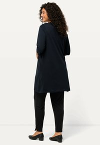 Marineblaue lange Tunika mit Dreiviertelärmeln und fließendem Design, kombiniert mit schwarzen schmal geschnittenen Hosen und schwarzen Absatzschuhen. Glatte Textur, minimale Details.