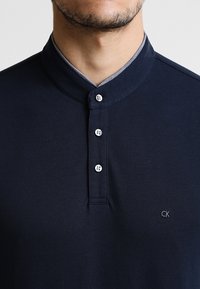 Polo de color navy con un acabado texturizado, que presenta un cuello con solapa, cuatro botones y un sutil logo de CK en el lado izquierdo.
