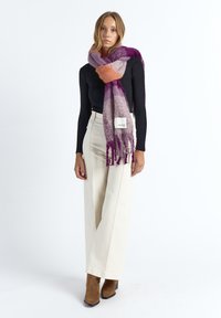 Multicolored wool scarf featuring purple, orange, and gray hues, with fringed ends. Worn casually over a black top with cream pants.