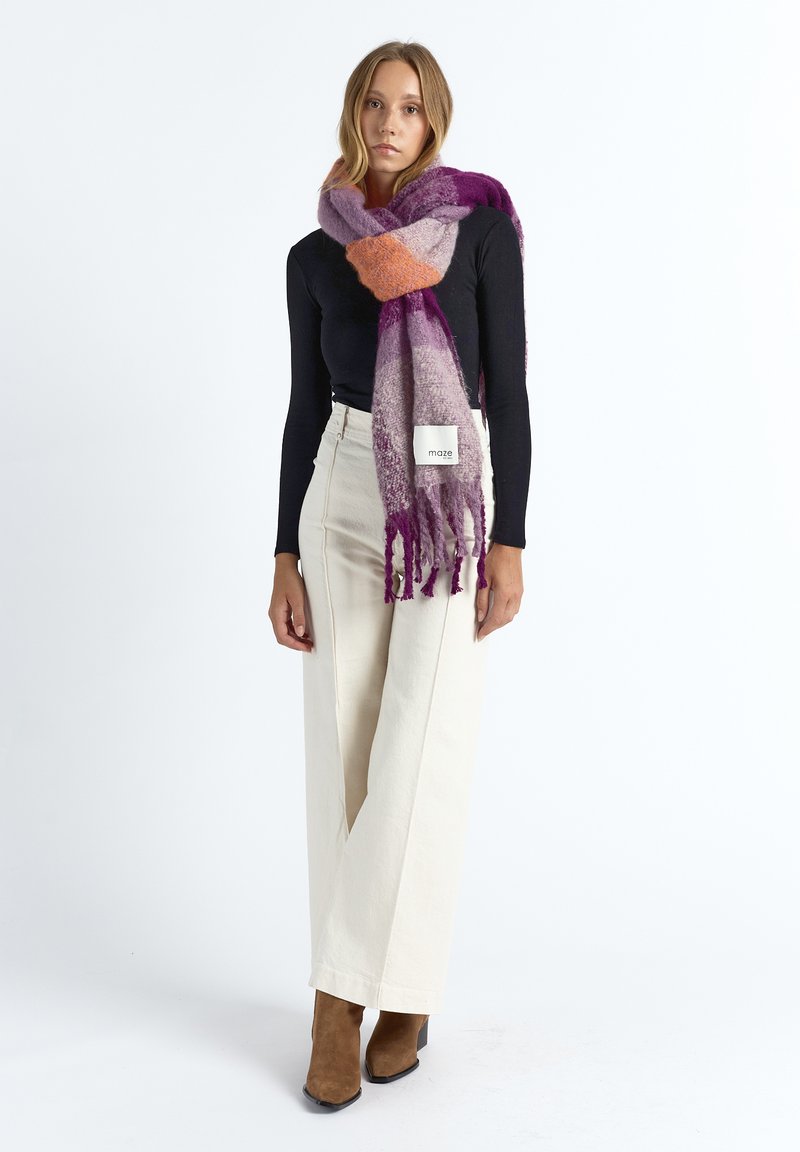 Multicolored wool scarf featuring purple, orange, and gray hues, with fringed ends. Worn casually over a black top with cream pants.