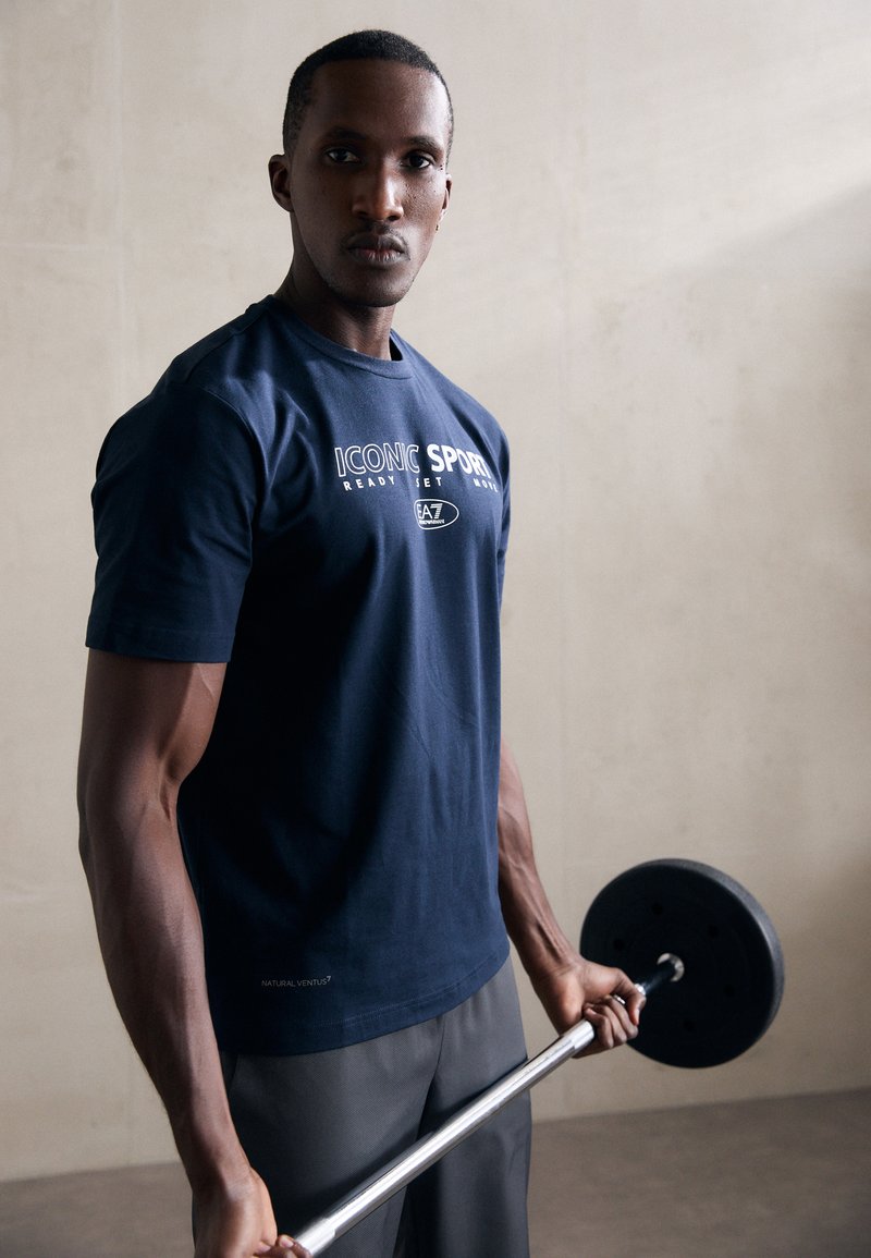 T-shirt de algodão azul marinho com "ICONIC SPORT" impresso em branco, com mangas curtas e decote redondo, usado por uma pessoa segurando uma barra de pesos.