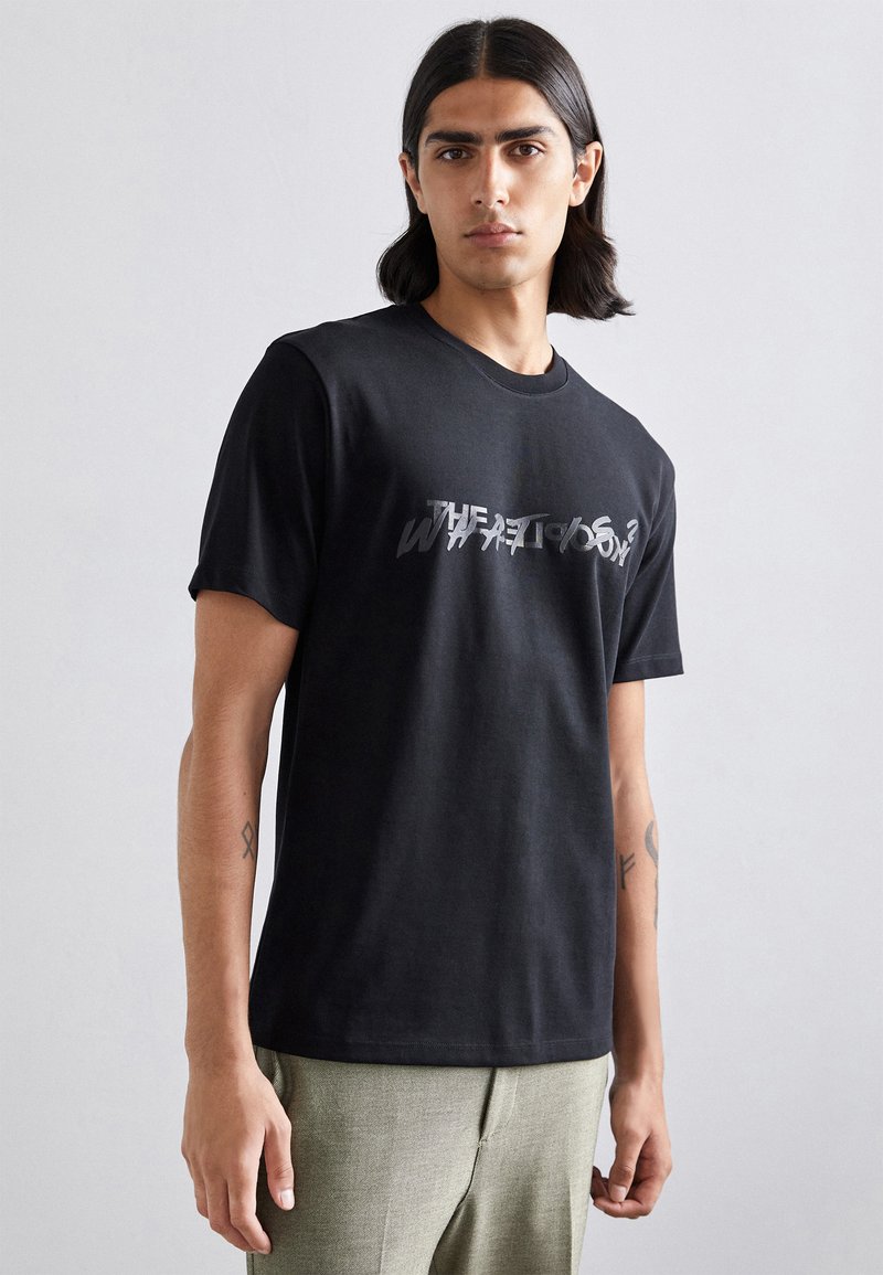 Black cotton t-shirt with a round neckline, featuring a distressed graphic print in silver text on the front. Short sleeves and relaxed fit.