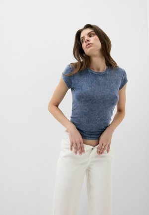 Frau in einem blauen kurzärmeligen Hemd und weißen Hosen mit Händen nahe der Taille, die leicht nach oben und seitlich blickt vor einem einfarbigen weißen Hintergrund.
