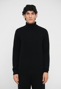 Schwarzer Strickrollkragenpullover mit glatter Textur, lässiger Passform und gerippten Bündchen. Modell trägt ihn vor einem schlichten weißen Hintergrund.