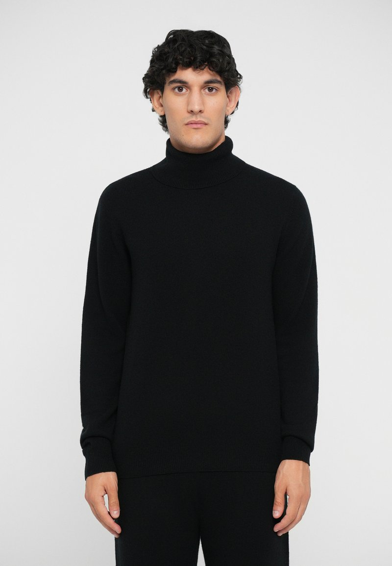 Schwarzer Strickrollkragenpullover mit glatter Textur, lässiger Passform und gerippten Bündchen. Modell trägt ihn vor einem schlichten weißen Hintergrund.