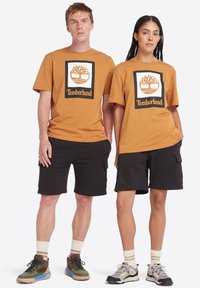 Camisetas de algodón marrón con el logotipo de Timberland en blanco y negro, combinadas con pantalones cortos negros. El calzado presenta colores apagados y suelas texturizadas.
