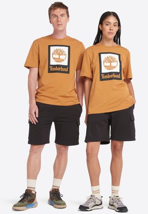 Braune Baumwoll-T-Shirts mit schwarzem und weißem Timberland-Logo, kombiniert mit schwarzen Shorts. Die Schuhe haben dezente Farben und strukturierte Sohlen.