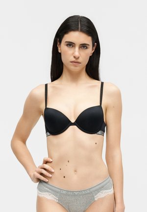 Γυναίκα με μακριά σκούρα μαλλιά, φορώντας μαύρο σουτιέν και γκρι εσώρουχο με δαντελένιες λεπτομέρειες, όρθια με το ένα χέρι στη μέση της μπροστά από απλό φόντο.