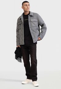 Prošívaná šedá bunda s límcem, dvěma kapsami na hrudi, nošená přes černé tričko, doplněná tmavými džíny a bílými teniskami.