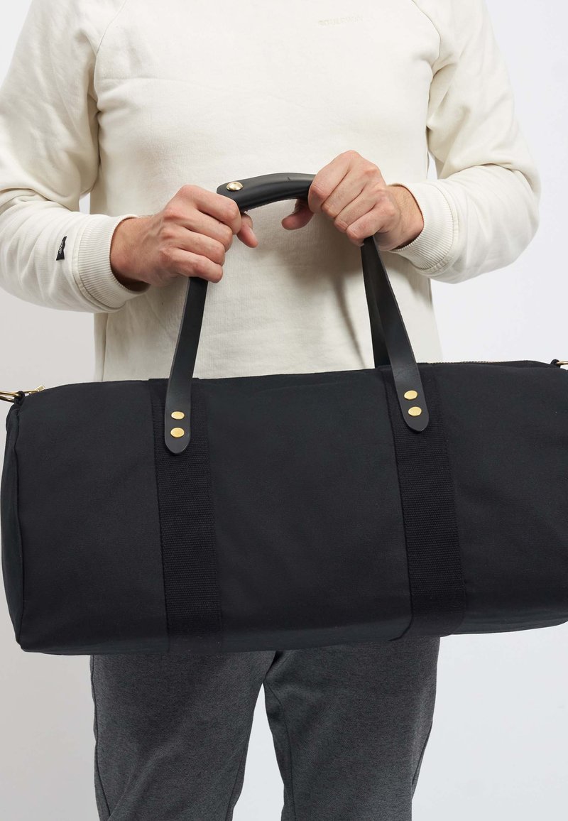 Černý sportovní vak s pevnými černými koženými uchy, zlatými kovovými detaily a texturovaným tkaninovým tělem. Minimalistický design, obdélníkový tvar.