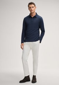 Polo a maniche lunghe blu navy con colletto classico, abbinato a pantaloni bianchi e stivali scuri in suede, con un sottile logo sul petto.