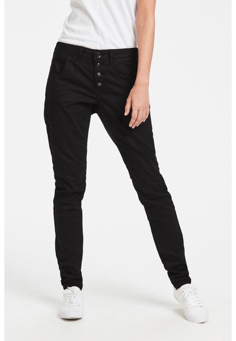 Personne portant un pantalon slim noir à fermeture quatre boutons et des baskets blanches, debout avec une jambe légèrement pliée.