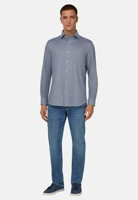 Mann steht frontal, trägt ein hellblaues Hemd mit Knöpfen, blaue Jeans und dunkelblaue Turnschuhe, vor einem schlichten Hintergrund.