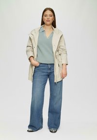 Hellbeige wasserdichte Jacke mit verstellbarem Kapuzen, über einem blauen, gerippten Oberteil und weiten blauen Jeans, mit silbernen Hardware-Details.