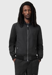 Czarna kurtka bomberka wykonana z gładkiego materiału, z zamkiem błyskawicznym z przodu, prążkowanymi mankietami i hemem, oraz dwiema kieszeniami po bokach. Dyskretny logo na klatce piersiowej.