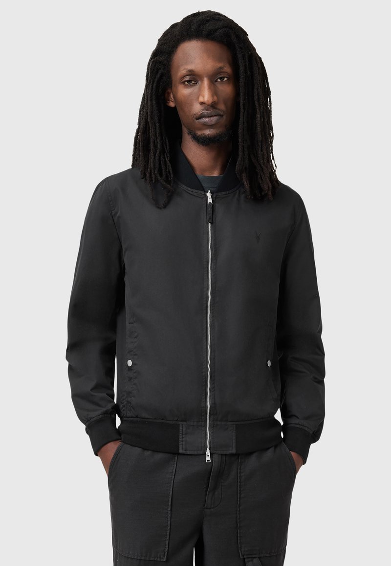 Czarna kurtka bomberka wykonana z gładkiego materiału, z zamkiem błyskawicznym z przodu, prążkowanymi mankietami i hemem, oraz dwiema kieszeniami po bokach. Dyskretny logo na klatce piersiowej.