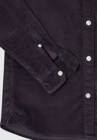 Ciemnofioletowa koszula z korduroy z mankietem na guziki, białymi guzikami oraz teksturowanym, prążkowanym materiałem i zaokrąglonym dołem.