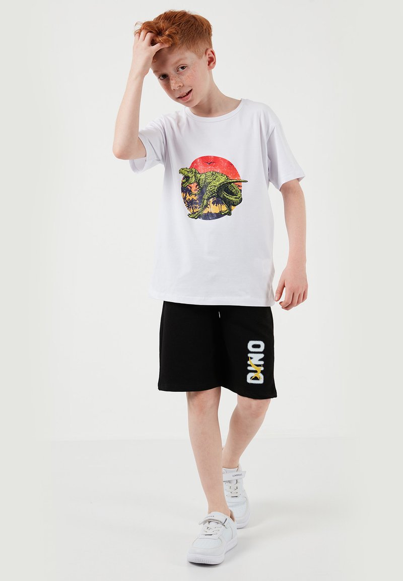 Garçon aux cheveux roux portant un T-shirt blanc avec un motif de dinosaure vert, un short noir avec le texte "DINO" et des baskets blanches, marchant vers l'avant à l'intérieur.