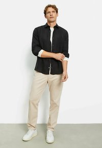 Schwarzes Hemd mit Knopfleiste über einem weißen Rundhalsausschnitt, kombiniert mit hellbeigen Hosen und weißen Sneakers. Schlichtes, strukturiertes Design.