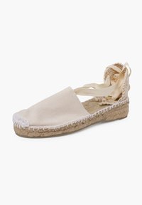 Espadrilla beige chiaro con tomaia in tela, suola in giuta e cuciture bianche. Presenta una morbida cinturina legata alla caviglia e un design con il tallone scoperto.
