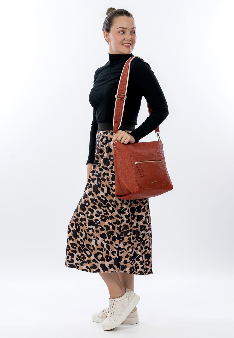 Bruine leren schoudertas met een gestructureerd oppervlak, gouden ritssluiting en een patroonriem. Model draagt een zwart topje en een rok met luipaardprint.