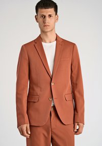 Americana de terracota con un diseño de un solo botón, dos bolsillos delanteros y botones marrones, combinada con una camiseta blanca de cuello redondo.