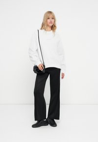 Valge suur suurus sweatshirt, mustad laiad treeningpüksid külgne erineva triibuga ja mustad tossud. Mudel hoiab käes väikest musta rätsepikkutakki.