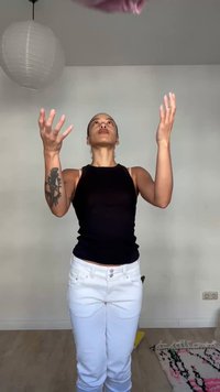 Femme en haut noir sans manches et pantalon blanc lançant un objet rose vers le haut à l'intérieur, debout près d'une lampe blanche suspendue et d'un tapis à motifs.