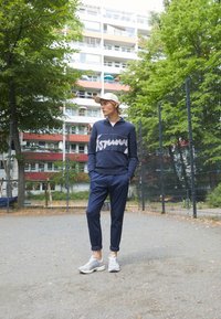 Sudadera navy con cremallera y texto gráfico blanco, combinada con joggers taperados navy y zapatillas gris claro. Posicionada al aire libre entre vegetación.