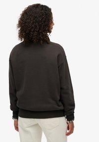 Donkerbruine, oversized trui met geribbelde boorden en zoom, met een zachte textuur. Een geruit overhemd steekt zichtbaar onder de mouwen vandaan.