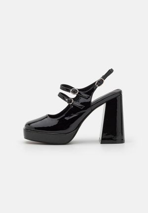 Talons plats en cuir verni noir avec un bloc épais, dotés de deux sangles réglables avec des accents en métal argenté rond.