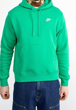 Mężczyzna w jasnozielonej bluzie Nike z kapturem, przednią kieszenią i troczkami, w połączeniu z ciemnoniebieskimi dżinsami na białym tle.