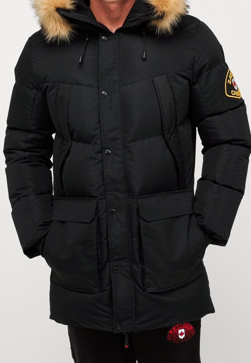 Veste noire matelassée avec capuche bordée de fourrure, boutons-pression et plusieurs poches avant ; comporte un écusson sur le bras gauche. Texture douce.