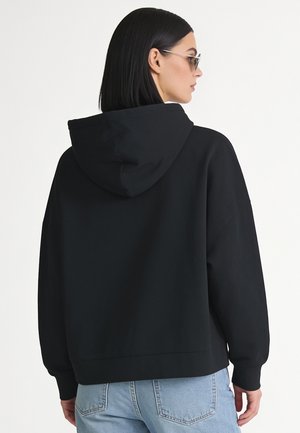 Femme aux cheveux noirs et lisses portant des lunettes de soleil, un sweat à capuche noir et un jean bleu clair, vue de dos sur fond blanc.
