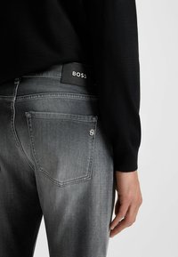 Jeans in denim grigio scuro con un leggero sbiadimento, dotati di tasche posteriori e un'etichetta nera con il marchio "BOSS". Il materiale sembra avere una texture.