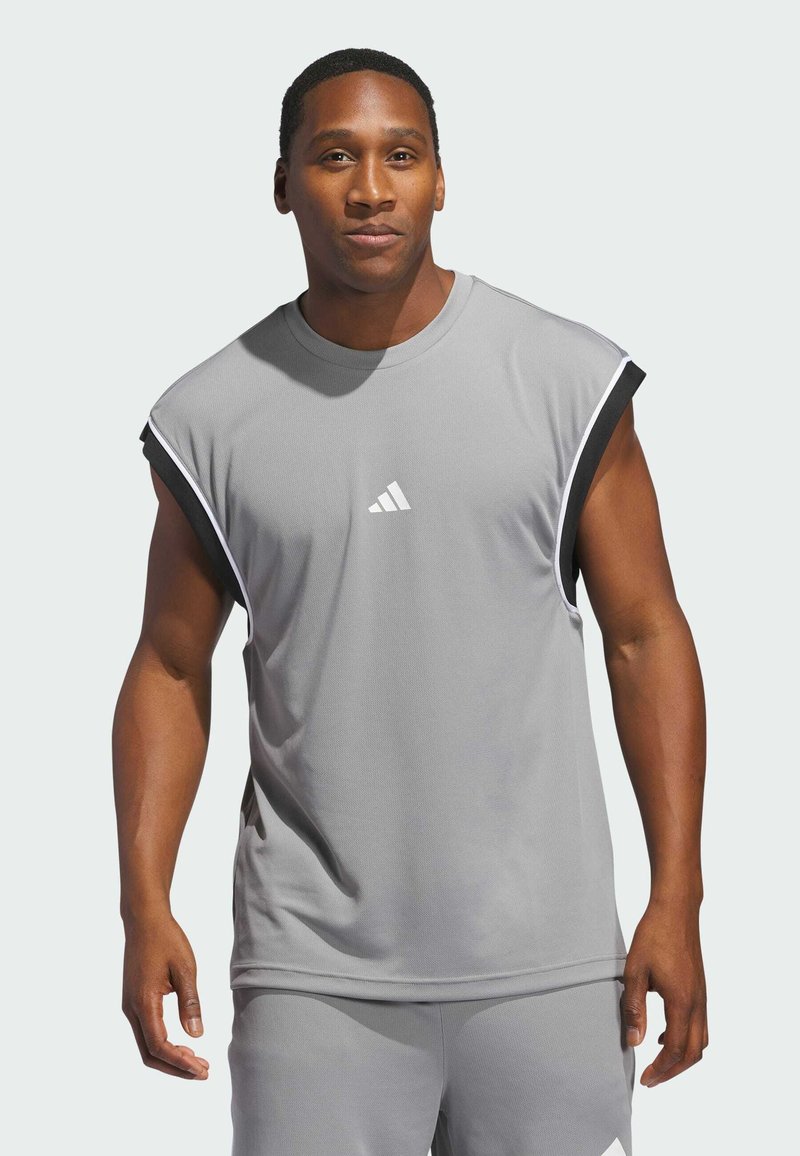 Hombre vistiendo una camiseta deportiva sin mangas de color gris y pantalones cortos con el logo de Adidas, de pie contra un fondo claro y liso.