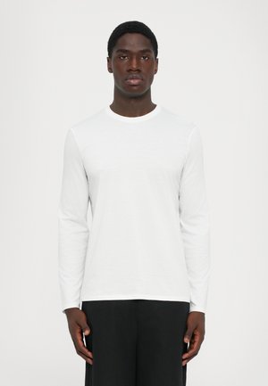 Young man with dark skin wearing a plain white long-sleeve shirt and black pants, standing against a light gray background.
