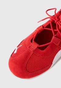 Scarpa sportiva rossa realizzata in materiali mesh e suede, con punta rotonda, lacci e accenti bianchi ai lati.