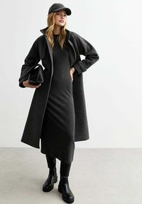 Donkergrijze lange jas over een geribbelde jurk, zwarte enkellaarsjes en een pet. De outfit omvat een textuurachtige zwarte handtas voor extra detail.
