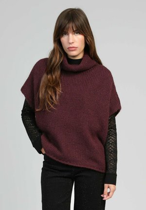 Femme aux longs cheveux bruns portant un poncho en maille maroon ample par-dessus un top en dentelle noire à manches longues et un pantalon noir avec de petites embellissements.