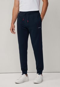 Marineblaue Sweatpants mit verstellbarem Bund, gerippten Bündchen und einem kleinen roten Logo am linken Oberschenkel. Kombiniert mit weißen Sneakers.