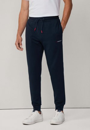 Marina blå sweatpants med dragsko i midjan, ribbade muddar och en liten röd logotyp på vänster lår. Tillsammans med vita sneakers.