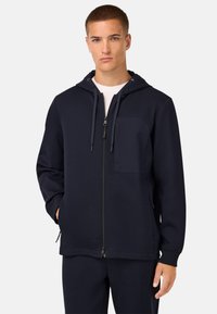 Mornarska zip-jakna iz mehkega blaga, z žepom spredaj, kapuco na vrvico in rebrastimi robovi. Gladka tekstura z minimalističnim dizajnom.