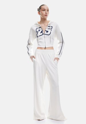 Witte cropped hoodie met een rits, voorzien van een marineblauw grafisch ontwerp en trekkoorden. Gecombineerd met witte wide-leg broeken, beide in zachte stof.