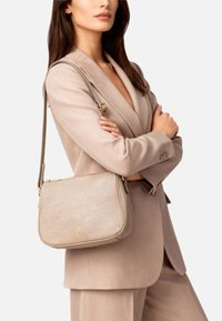Borsa a tracolla in pelle beige dalla forma arrotondata, con dettagli dorati e finitura testurizzata; indossata con una tracolla regolabile abbinata a un outfit neutro.