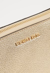 Złota torebka z teksturą, wyposażona w gładki zamek błyskawiczny oraz wytłoczoną nazwę marki "MICHAEL KORS" w złotym kolorze po boku.