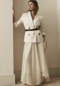 Femme en blazer beige à double boutonnage avec ceinture noire et longue jupe crème texturée se tenant à l'intérieur, s'appuyant sur un mur avec un fond neutre.