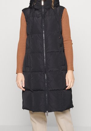 Veste noire matelassée sans manches avec fermeture éclair sur le devant, portée sur un haut marron à manches longues, pantalon beige et mains le long du corps.