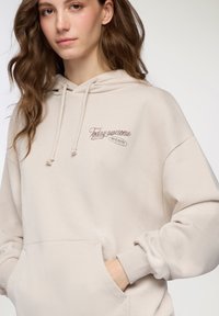 Beżowa bluza z kapturem, z sznurkiem, przednią kieszenią i ściągaczami. Zawiera haftowany tekst "Dziś wspaniale" i "BEAR MOTEL" w kolorze brązowym.