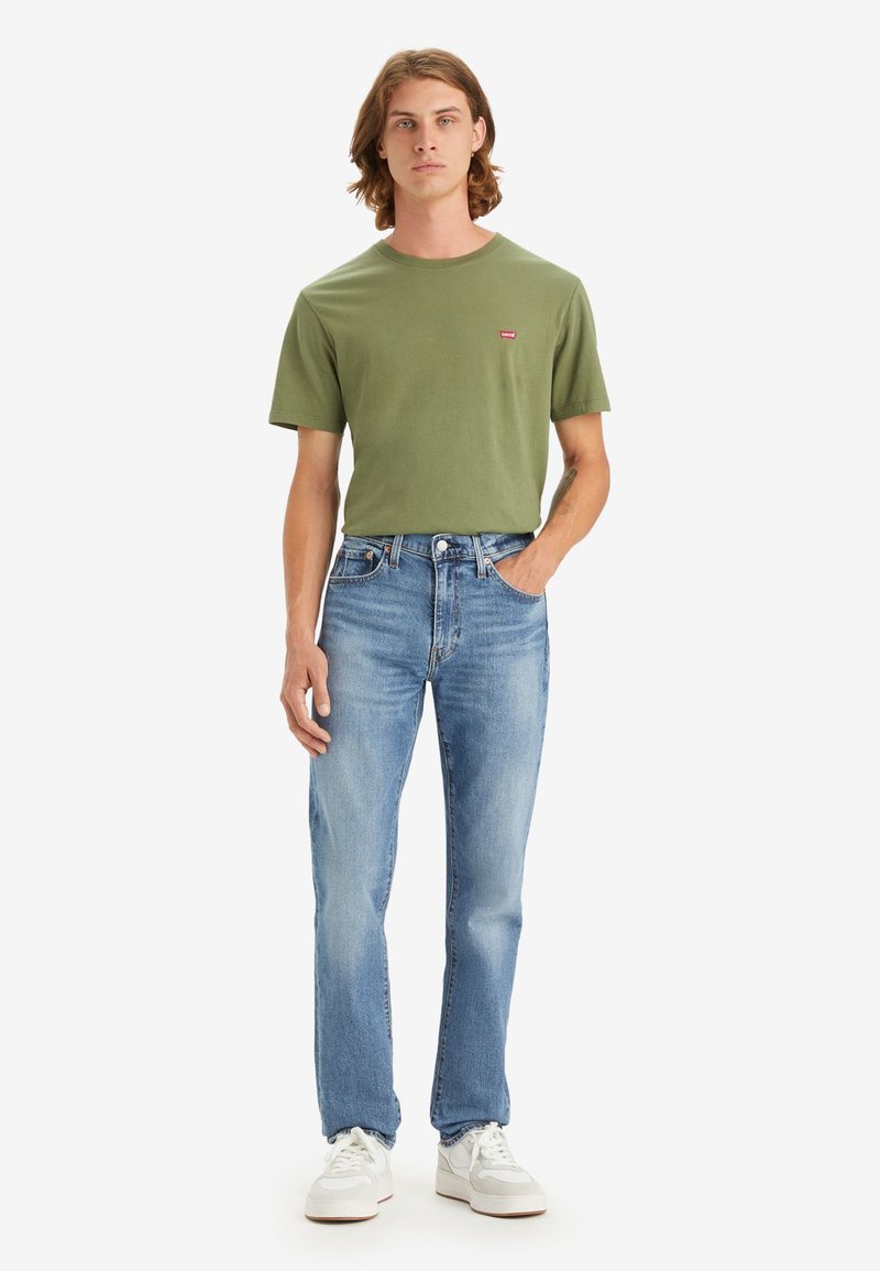 Camiseta de algodón color oliva con un pequeño logotipo rojo, combinada con jeans de mezclilla azul claro y zapatillas blancas. Ajuste y diseño simples.